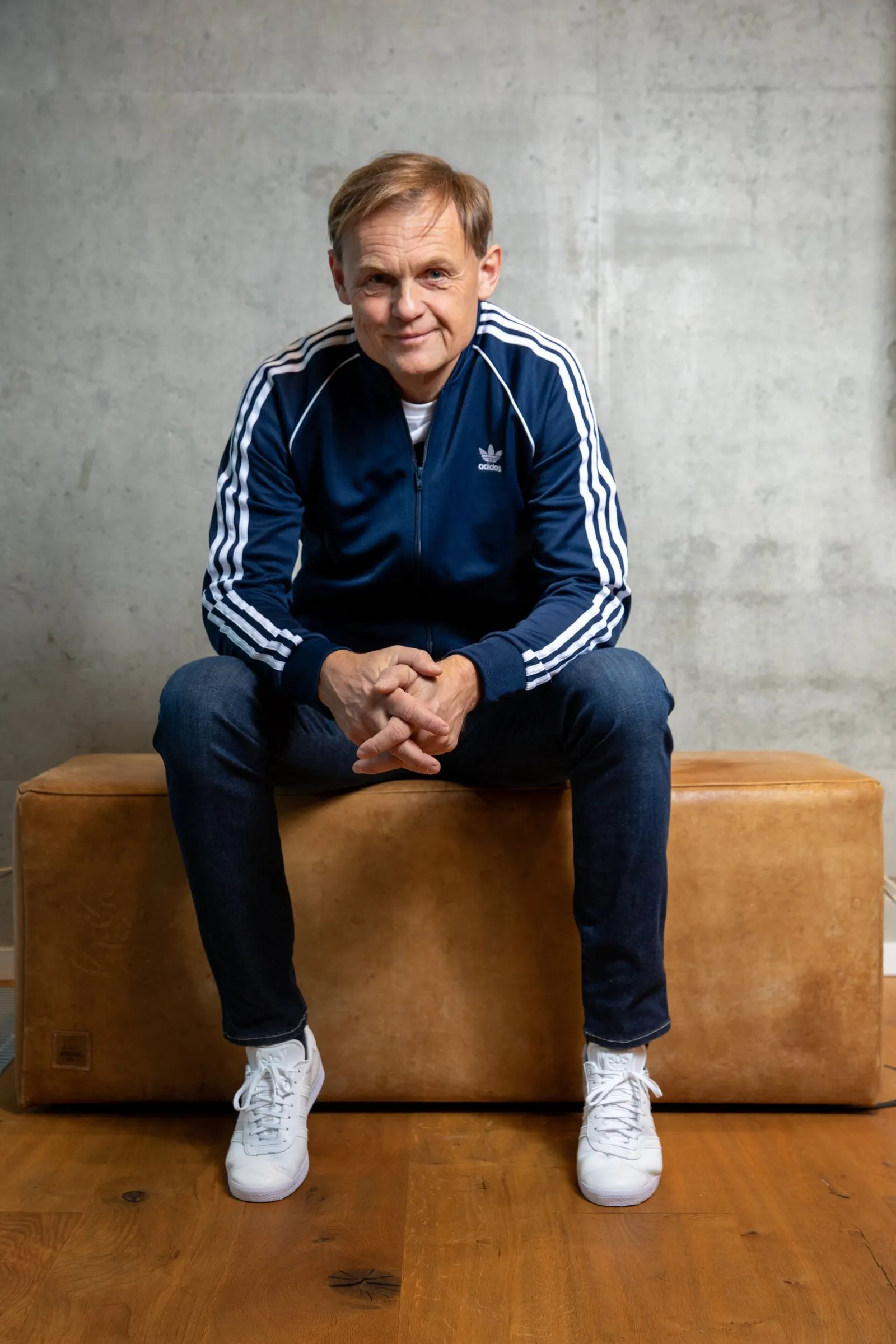 Adidas CEO Bjørn Gulden sits on a leather bench in a blue Adidas tracksuit.