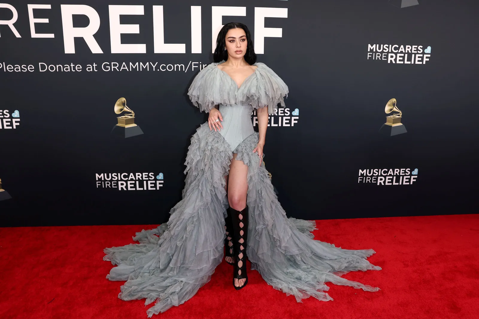 A person in a long, gray gown and high black boots stands on the red carpet.