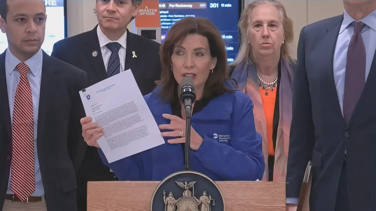 A woman in a blue jacket at a podium holding up a letter surrounded by several people.