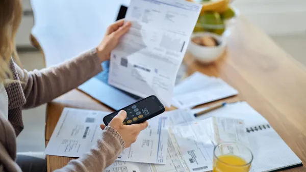 Woman using mobile phone while calculating the amount of her bills at home. Focus is on hand and cell phone.