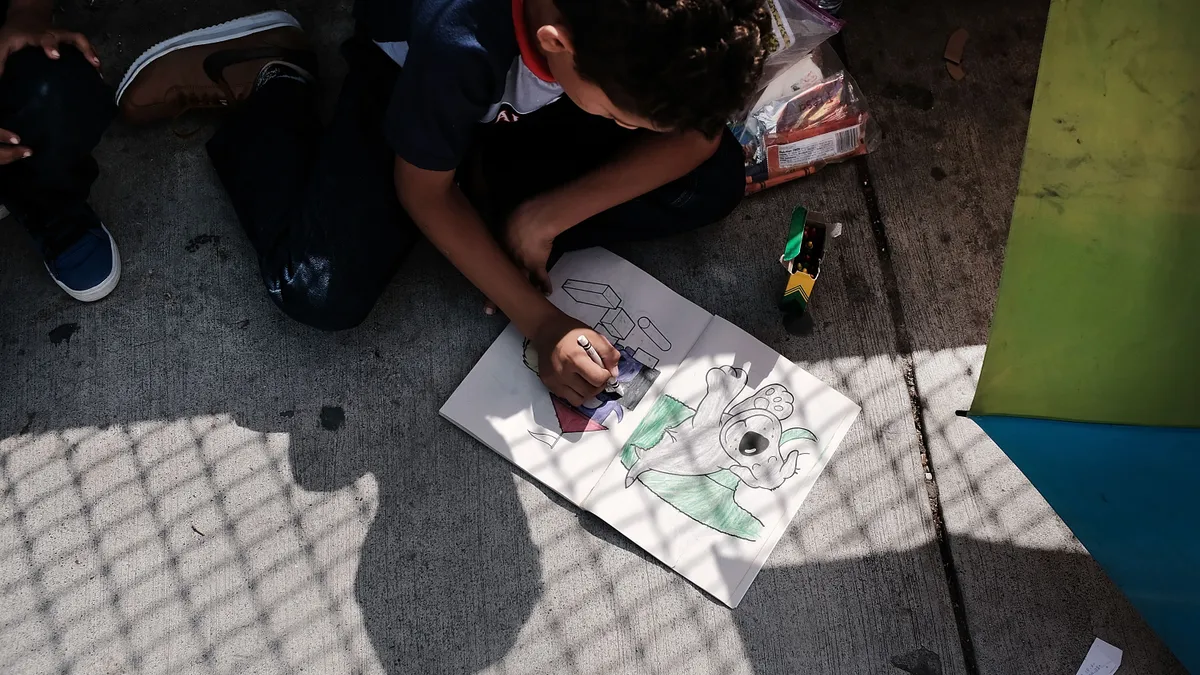 A child sits on concrete on the ground with head over a picture they are coloring with Crayons.