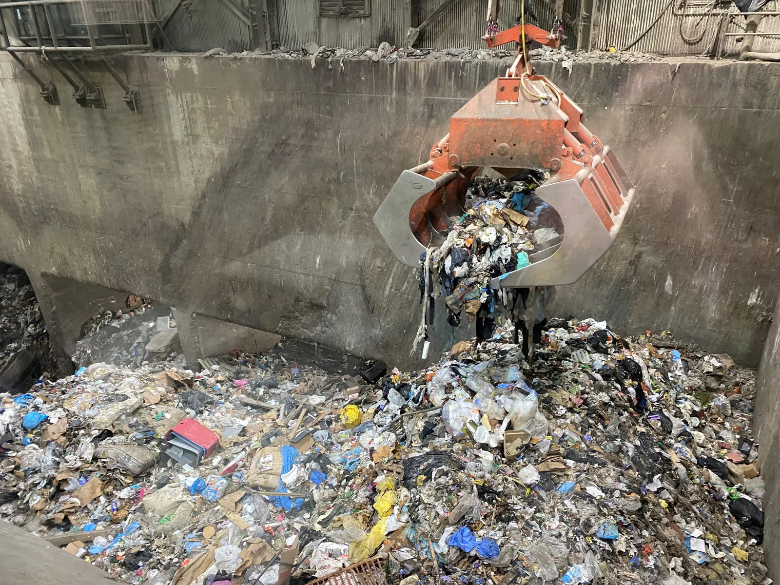 A large crane drops mixed trash into a large concrete pit in an enclosed building.