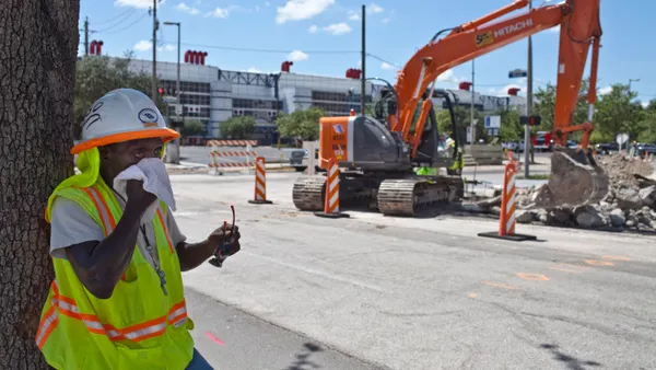 A construction worker in a white hard hat and yellow reflective vest wipes sweat from his face.