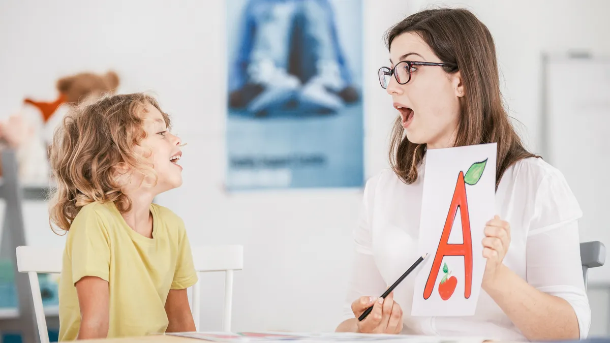 A student and adult sit across from each other at a desk. The adult holds a poster with the letter "A" and points to it while opening their mouth.