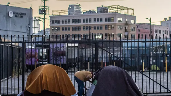 A person bends between two tents on a city street with a fence and buildings in the background.