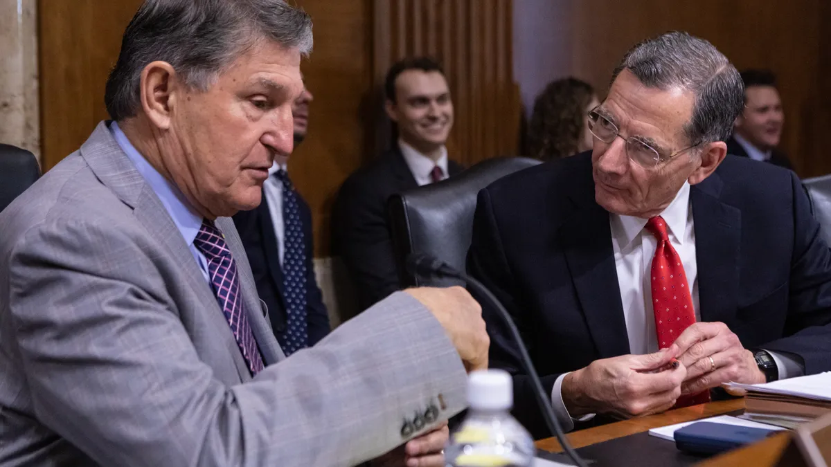 Two seated grey-haired men in suits and ties talk behind a wooden table with a man smiling in the background.