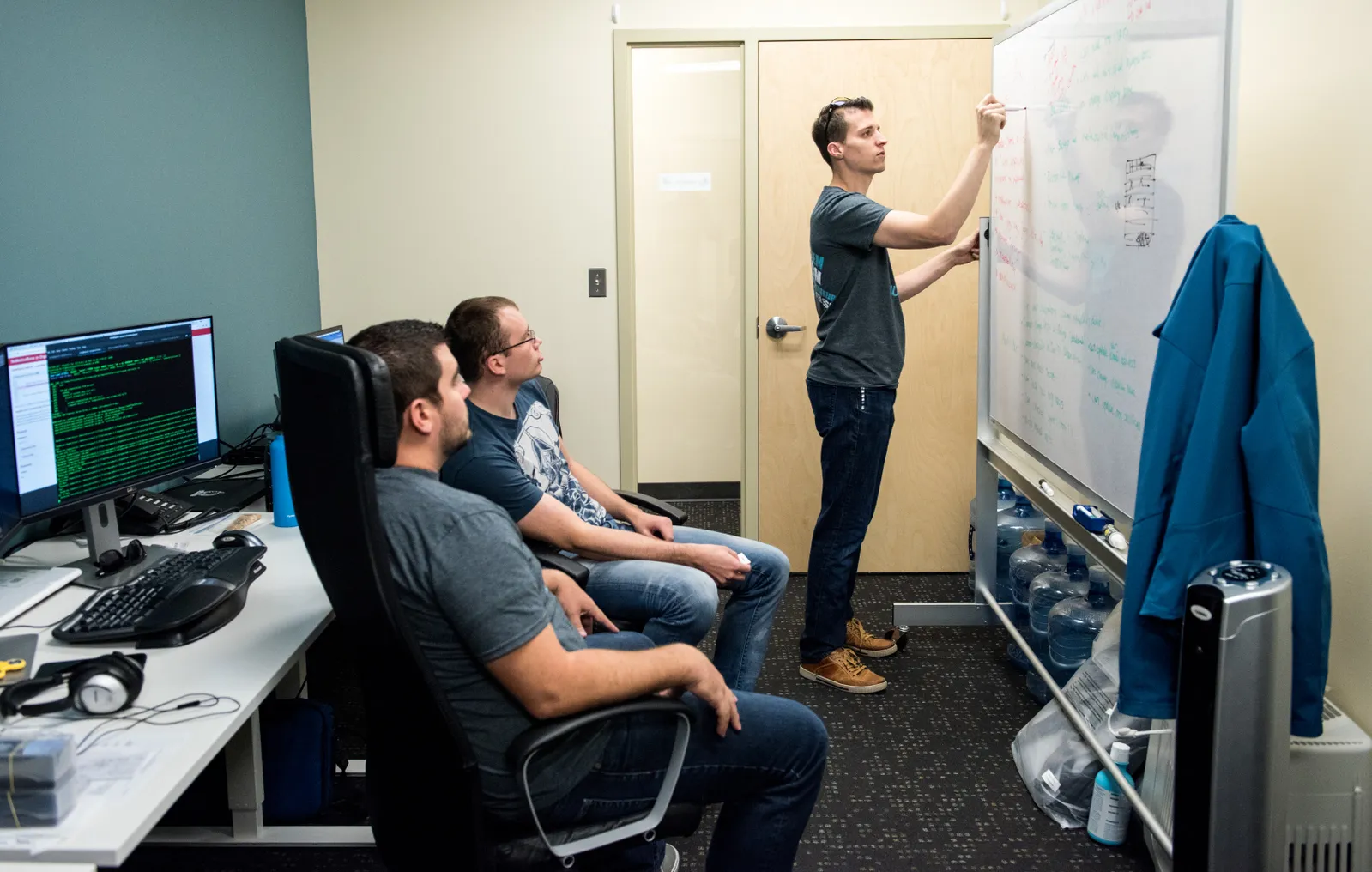 A person in a small room writes on a movable whiteboard as two people sitting in desk chairs look on. Behind them is a computer monitor with green code displayed.