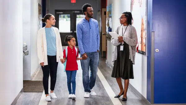 Three adults are standing in a school hallway. A child is holding on to two adults' hands and is wearing a backpack