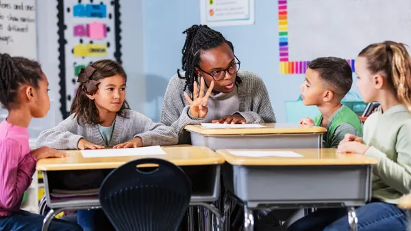An adult is sitting at a desk in a classroom with four students. The adult is holding up four fingers.