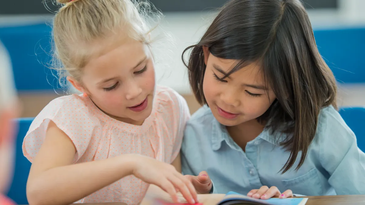 Two young students sit at a desk and look down and point at a book.