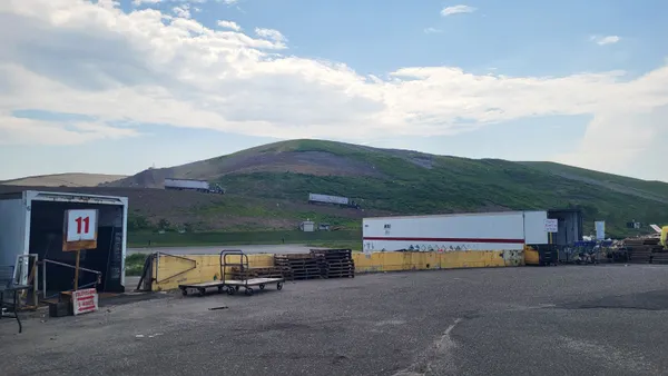 Several truck trailers and pallets sit on a concrete lot. Behind them, the mound of a landfill rises, with two 18-wheelers driving on a road along its slope.
