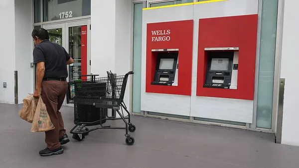 A person carrying bags with a shopping cart in tow walks past a Wells Fargo ATM at the front exterior of the bank's branch in Miami.