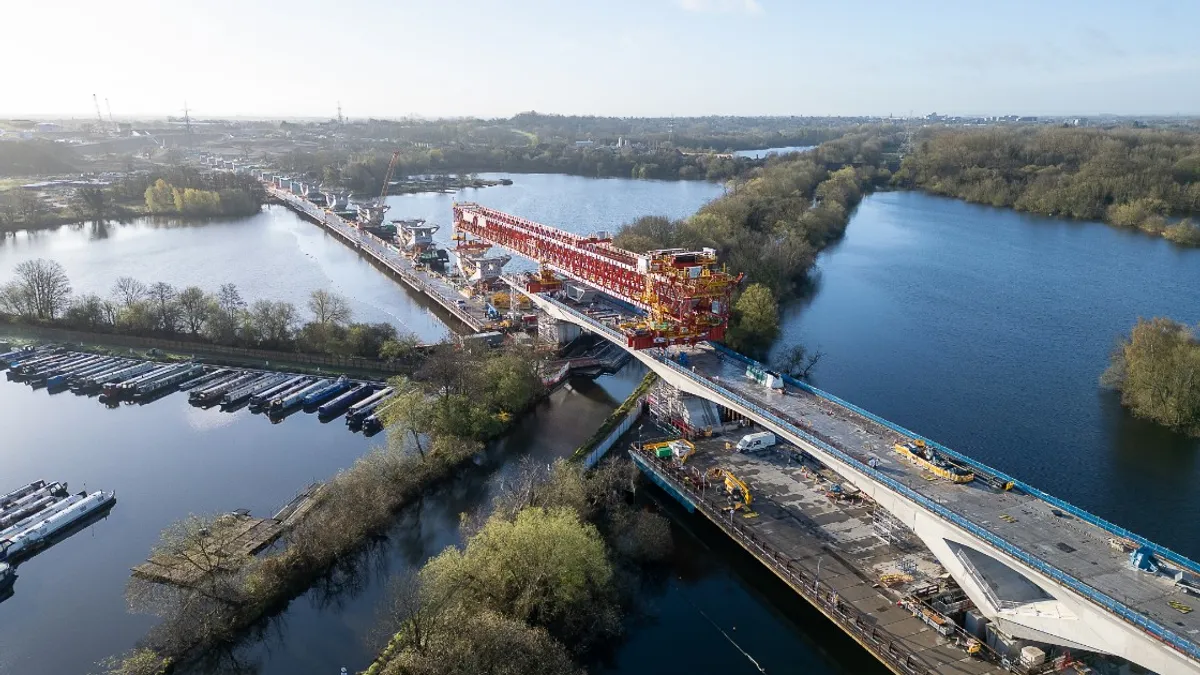 A large structure crosses a canal on a bright day. Boats, trees and blue water surround the construction.