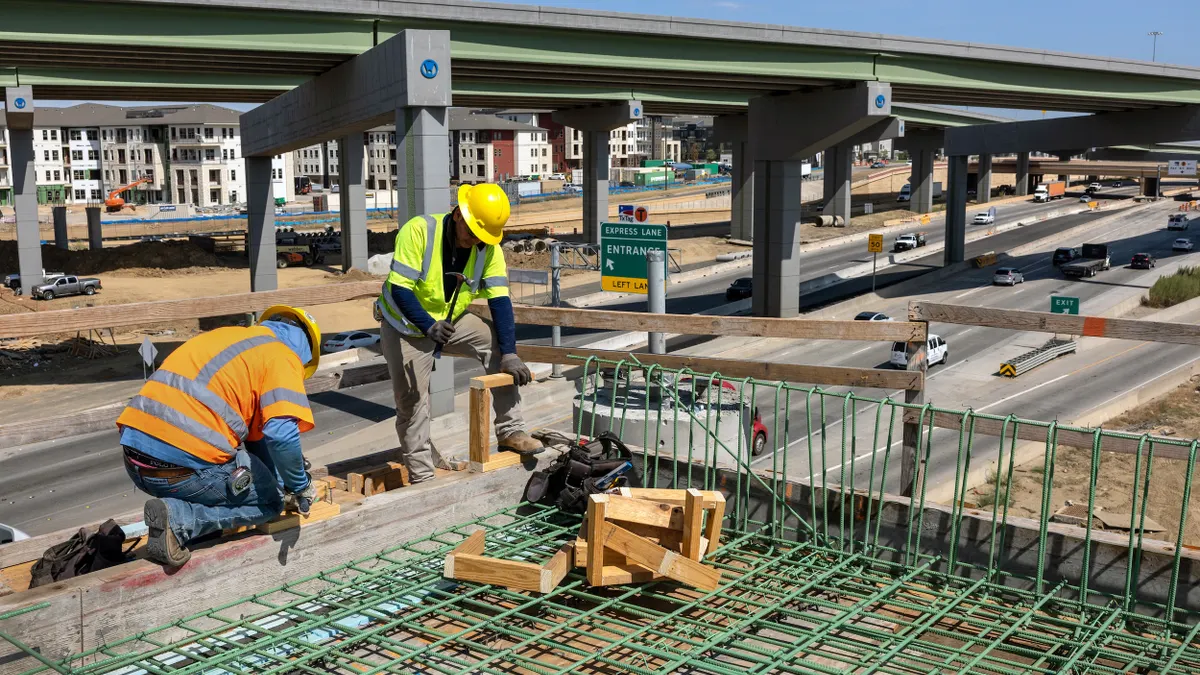 Two construction workers in hardhats with a highway behind them.