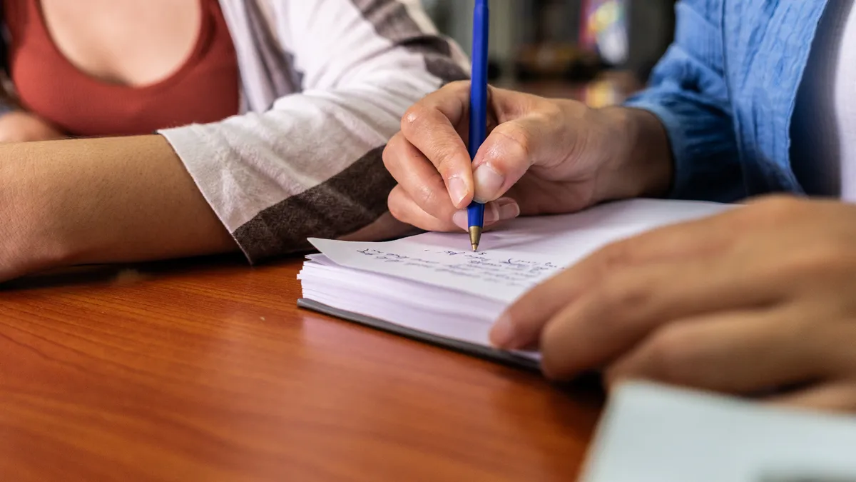 Closeup of the hands of two people sitting next to each other at a table. One is writing in a notebook with a pen.