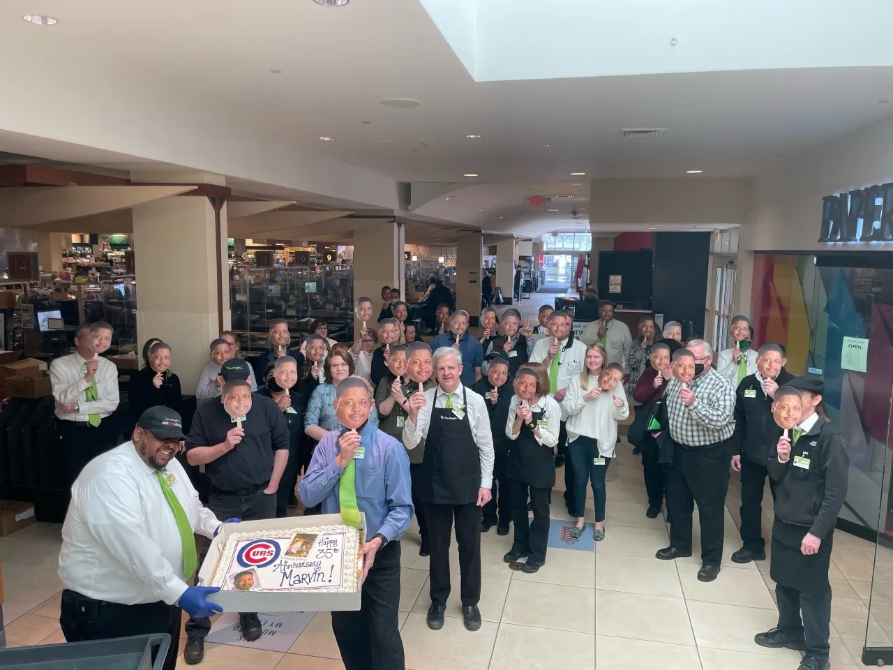 Two people hold a cake that says "Happy 35th anniversary Marvin" in front of dozens of people holding up cut-outs of the face of one of the people holding the cake.