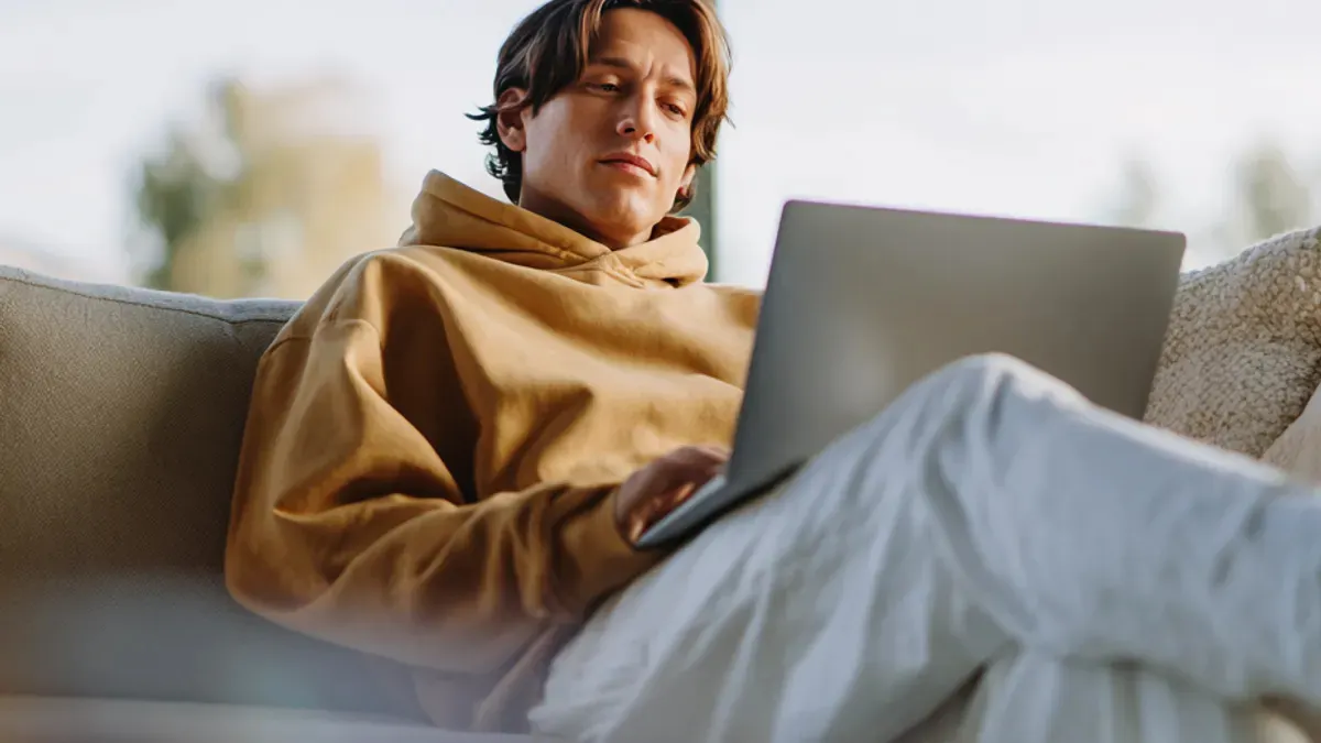 A person in a tan hoodie sits on a couch using a laptop, with natural light coming from a window behind them.