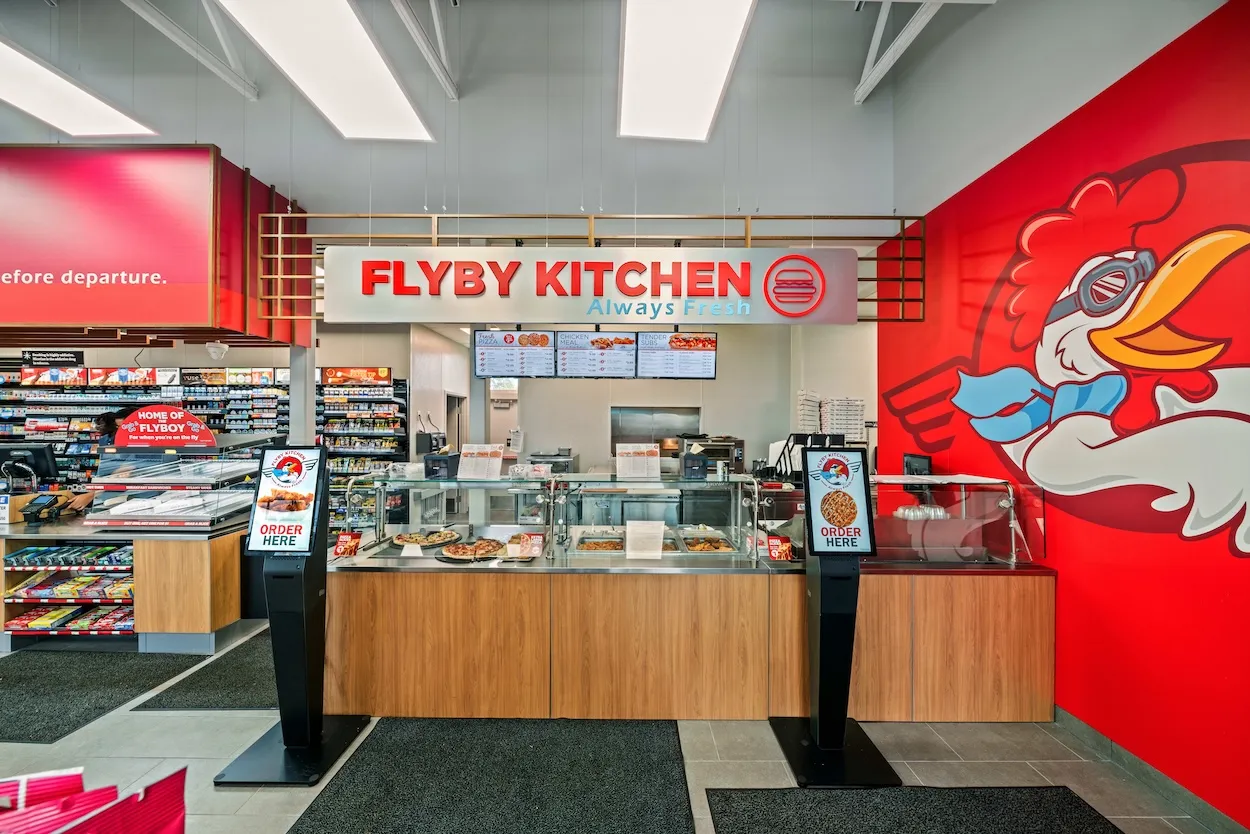 A photo of a foodservice counter in a store. A sign over the counter says &quot;Flyby Kitchen. Always fresh.&quot; there are two ordering kiosks and several glass-covered display areas. A logo on the wall to the right shows a chicken in aviation goggles and a scarf.