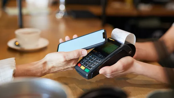 A person uses a smartphone for contactless payment at a café, holding it near a card reader. A coffee cup is visible in the background.