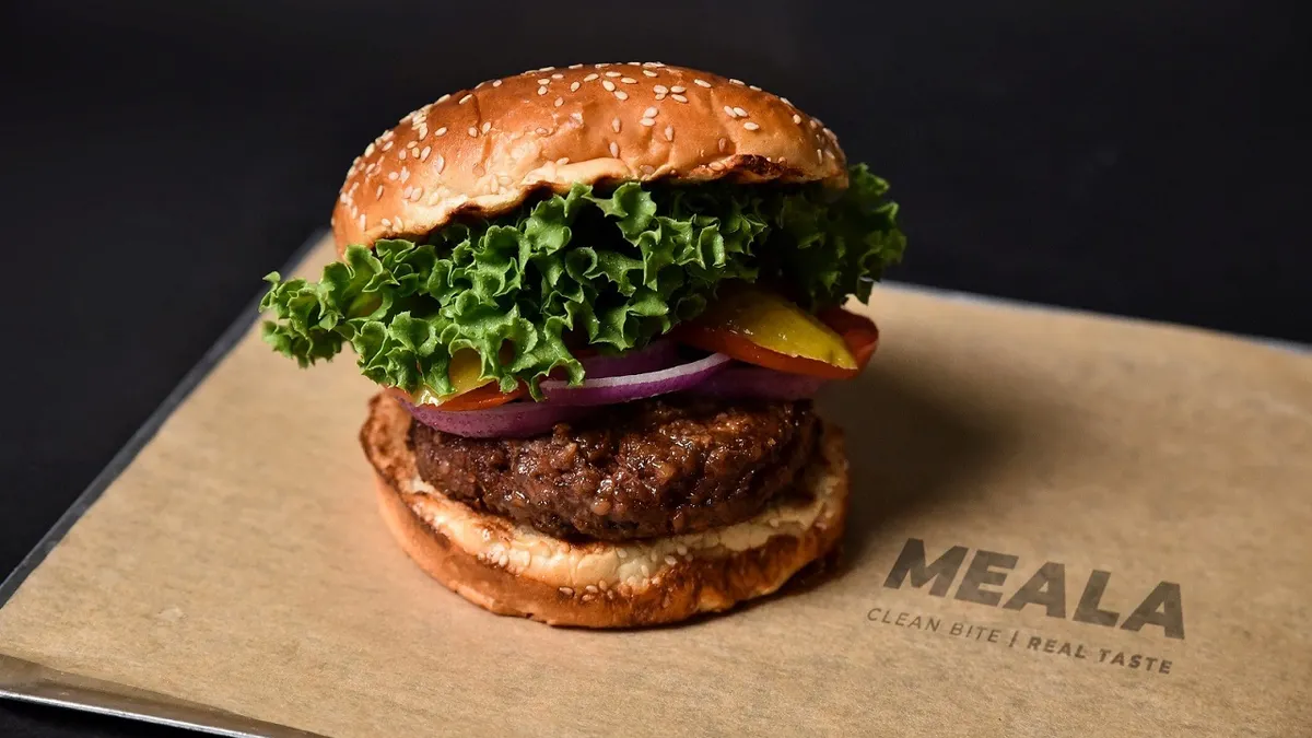 A small plant-based burger on a sesame seed bun with lettuce, tomatoes, red onions and cheese sits on a board featuring Meala's logo.