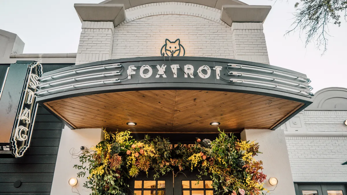 A photo of the exterior of a store. A neon sign on the canopy above the door reads "Foxtrot." A logo of a fox is on the wall above that, while a neon sign that says "Stag" is off to the left.