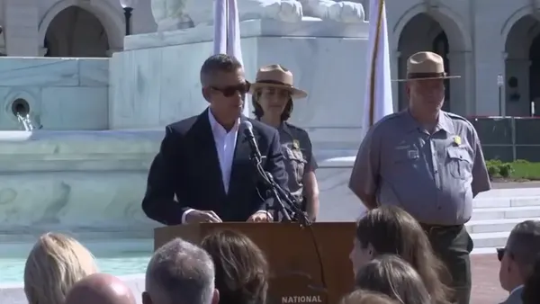 A man in a suit wearing sunglasses with two uniformed people alongside at a podium speaking to a group of people.