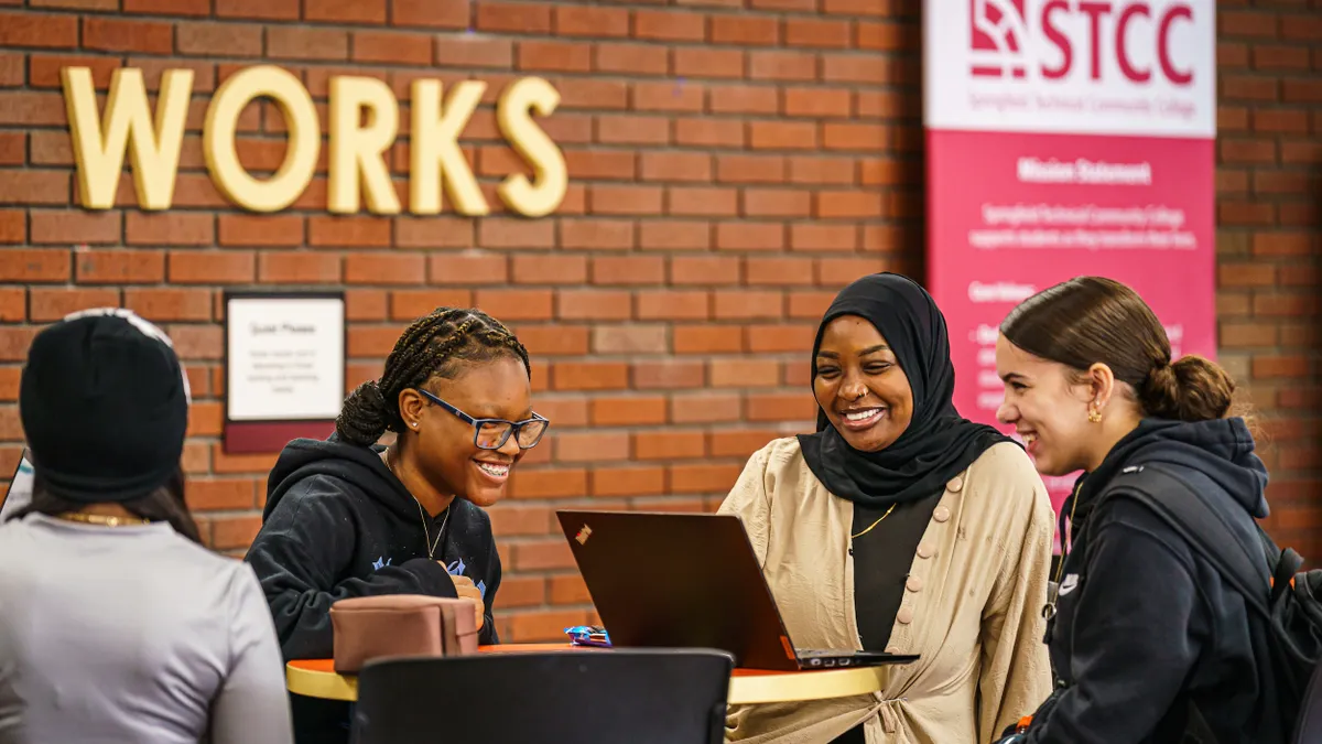 Three students are sitting at a table in a room with a red brick wall. There is an open laptop on the table. ANother person at the table has their back toward the camera