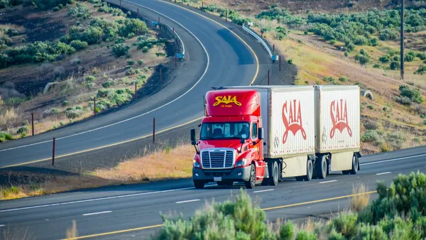 A Saia LTL semi-truck driving along a two-way highway with a hillside in the background.
