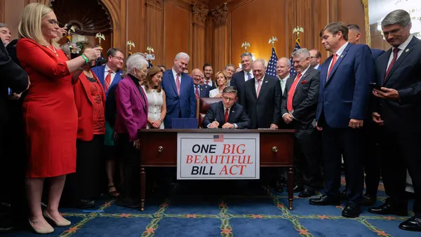 House Speaker Johnson signs a paper, while seated at a table with a sign that reads "One Big Beautiful Bill Act," while surrounded by a group of people.