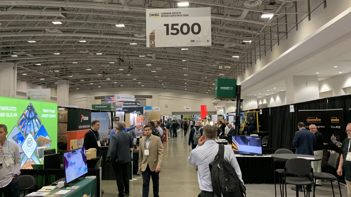 A wider shot of a gray-floored expo hall. Booths are set up along the edges of the photo, and a crowd of people are milling back and forth in the hall. All are dressed in business-casual attire.