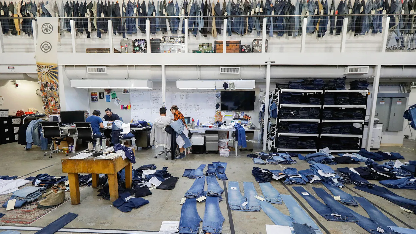 A spacious open studio featuring a lofted area with numerous jeans displayed in a row. Below the loft, there are two work desk areas occupied by several people. Jeans lay flat in the foreground.