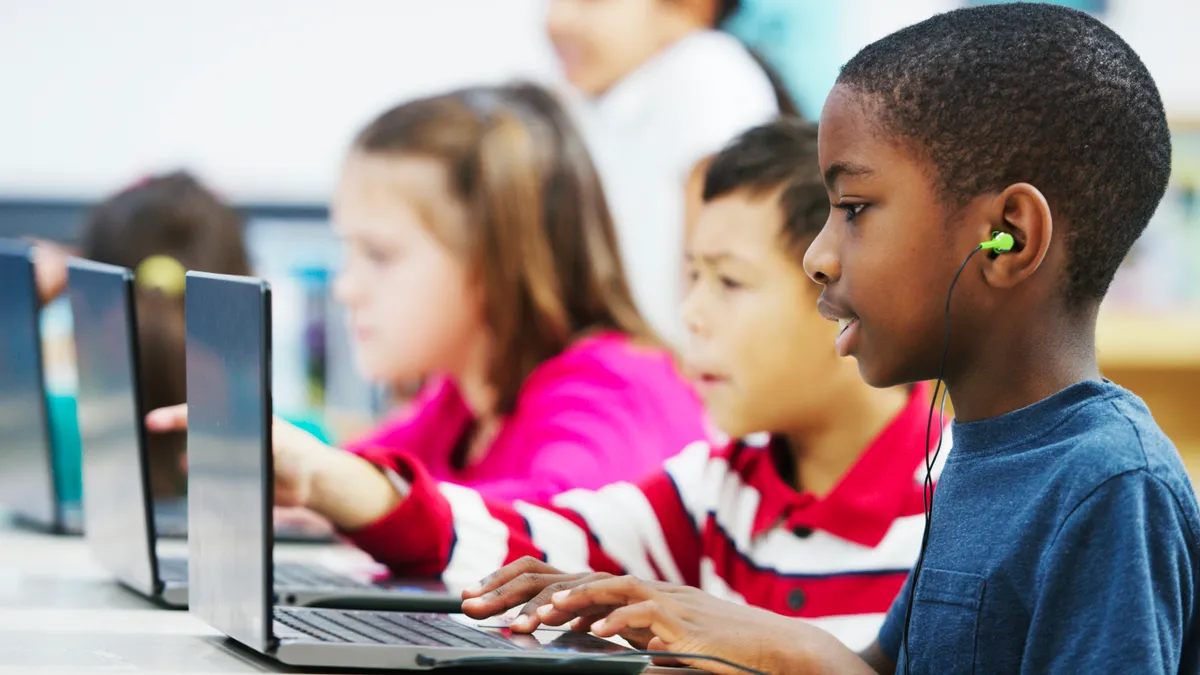 A handful of students are seated at a table with open laptops in front of them. The student closest to the camera is wearing headphones.