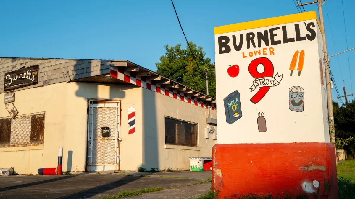 One-story cinder block building with sign in front that says "Burnell's Lower 9 Strong" and has pictures of a box that says "NOLA O's," a brown bag and a can of beans.
