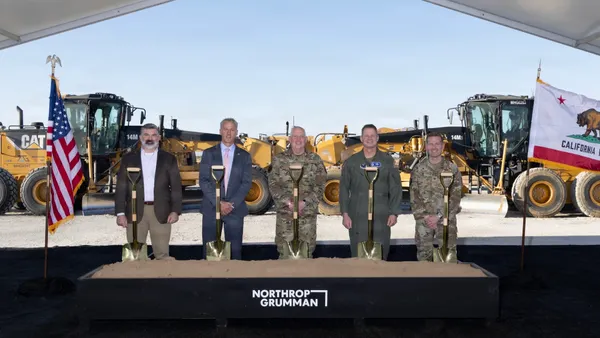 A group of people in suits and military fatigues stand in front of a black box featuring the name "Northrop Grumman."