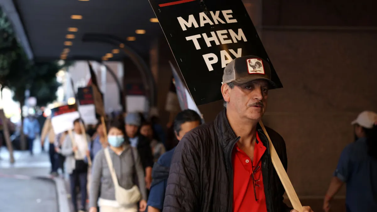 A worker on a picket line holds a sign reading "make them pay."