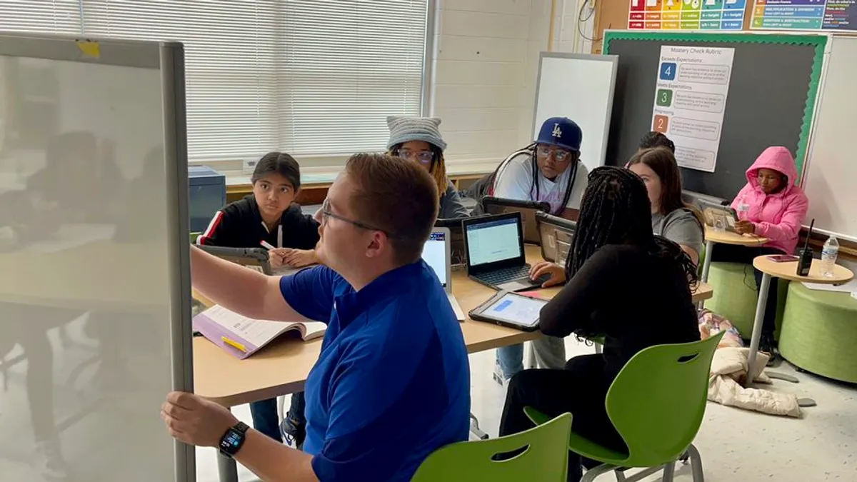 An adult and several students are seated at a table in a classroom. The teacher is writing on a white board and students are looking at the board or at computers on the table.s