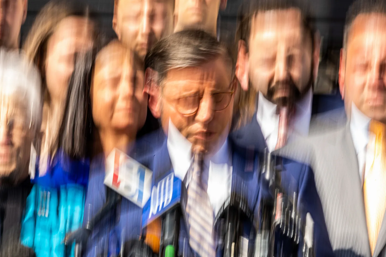 A group of people at a podium with a person dressed in business attire is focused in the front, the image has motion blur in a vertical direction.