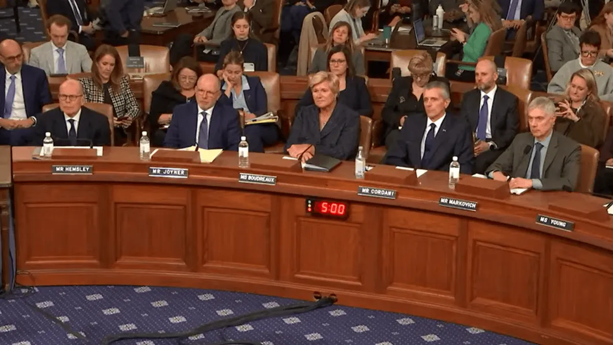 Six people in business clothes sit behind a dias in a congressional room.