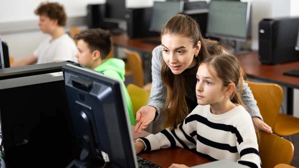 A teacher looks over a young student's shoulder as the student works at a computer.