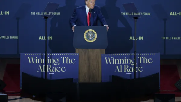 President Donald Trump stands at a lectern on a podium in front of a blue backdrop. Signs in front of him say "Winning the AI race" and the backdrop behind him says "The Hill & Valley Forum" and "All-In."