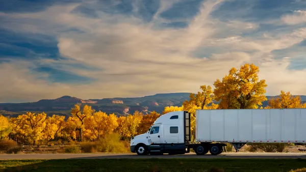 A tractor-trailer travels along a rural Colorado highway beneath a dramatic autumn sky with golden cottonwood trees in the background.