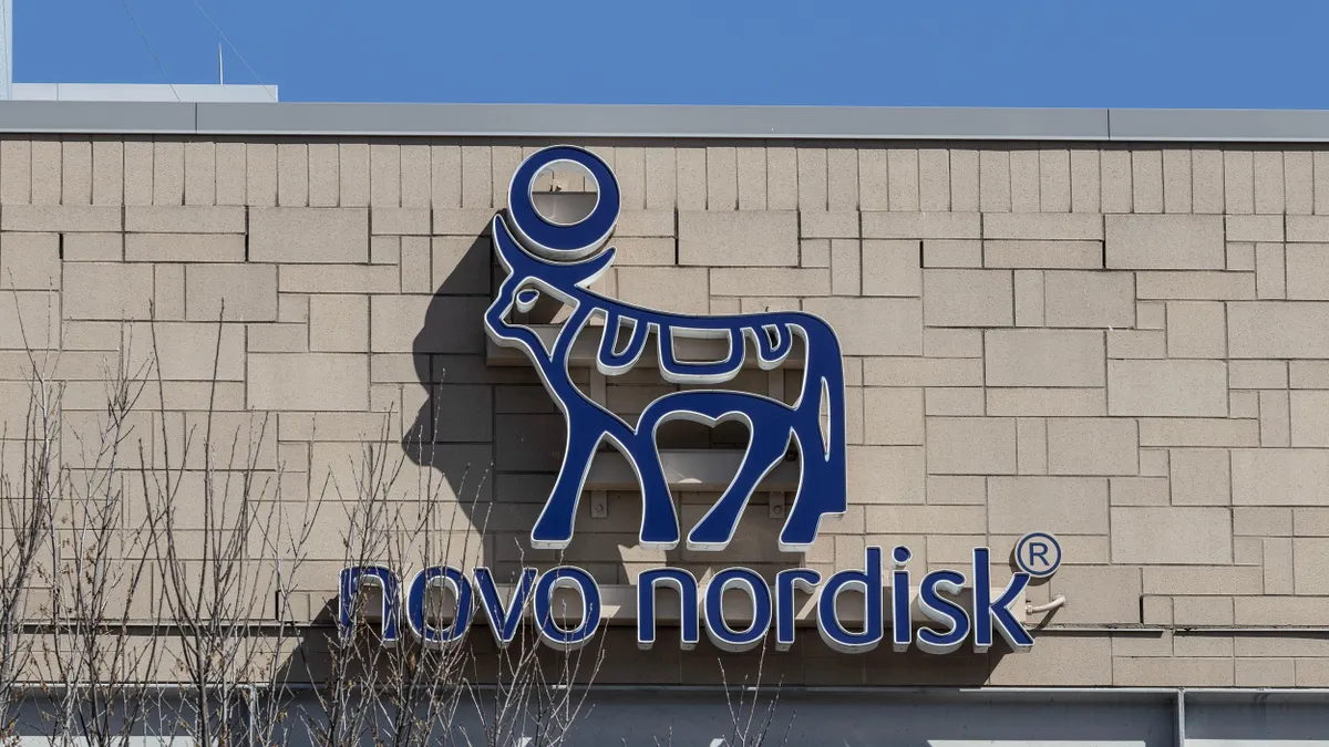 Close-up view of the Novo Nordisk logo mounted on the exterior wall of a building. The logo features a stylized blue Apis bull with a sun disk above its head, symbolizing the company's identity. The background shows a tan brick wall and part of a leafless tree in the foreground under a clear blue sky."