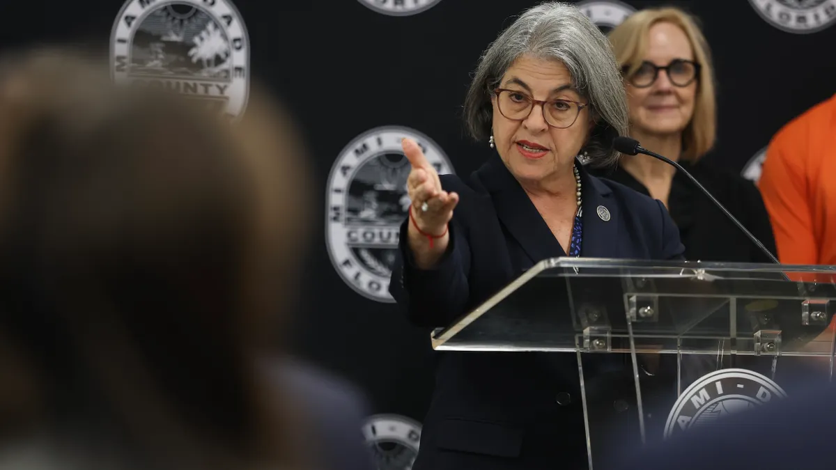 A person stands at a podium and gestures with one hand.