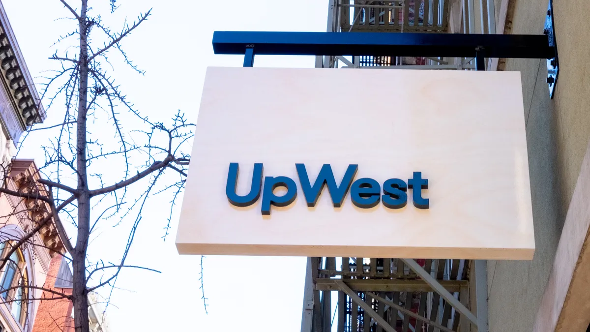 With tree branches and brick buildings to the left, a sign reading "UpWest" juts out from a building.