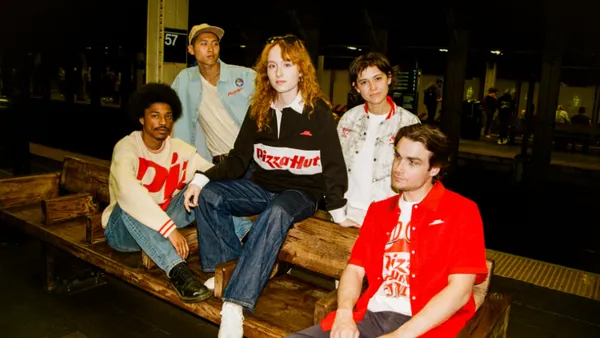 A group of five people wear Pizza Hut merchandise while sitting on and standing around a wood bench in an urban area.