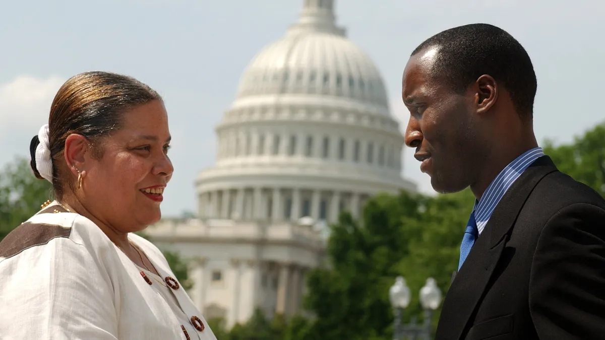 Two adults stand, facing each other outside. In the background is the dome of the U.S. Capitol in Washington, D.C.