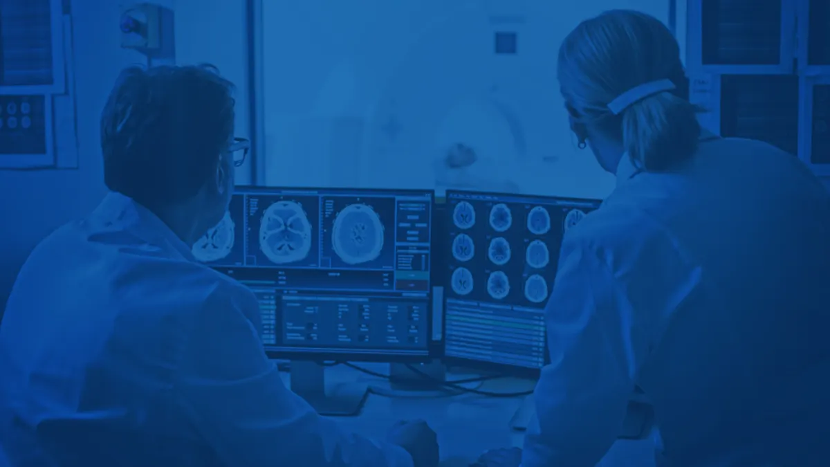 Two medical professionals in lab coats analyze brain scans on computer monitors while an MRI machine operates in the background.