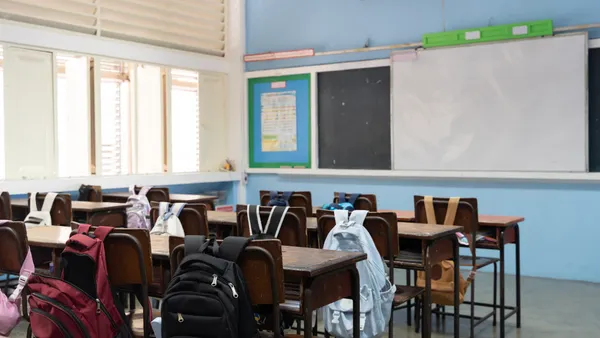 An empty classroom has backpacks rests on the back of chairs near desks.