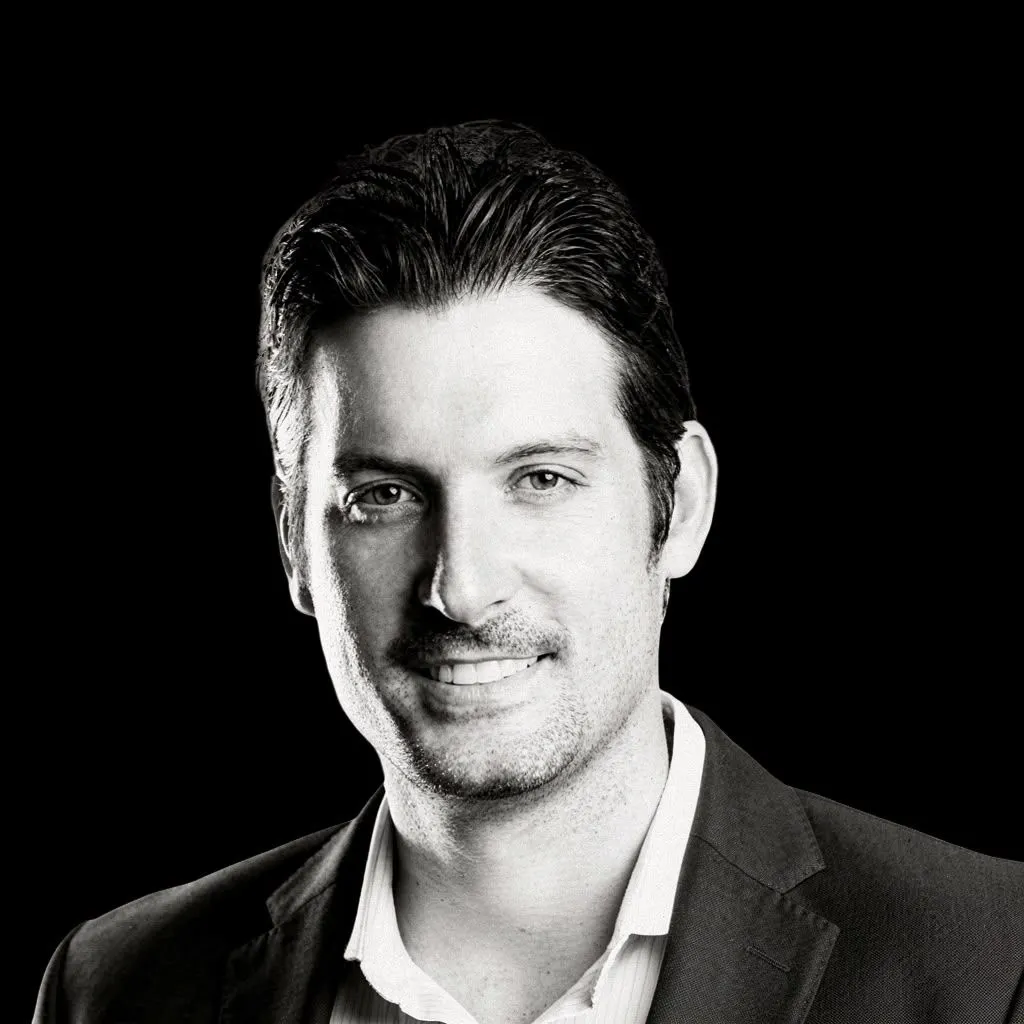 A black and white headshot of a person with slicked back hair and light goatee smiling.