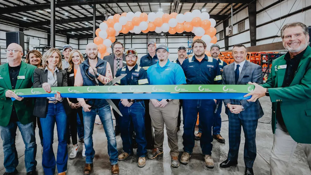 A group of people stand in a factory where they all smile behind a ribbon. One person in front holds scissors.